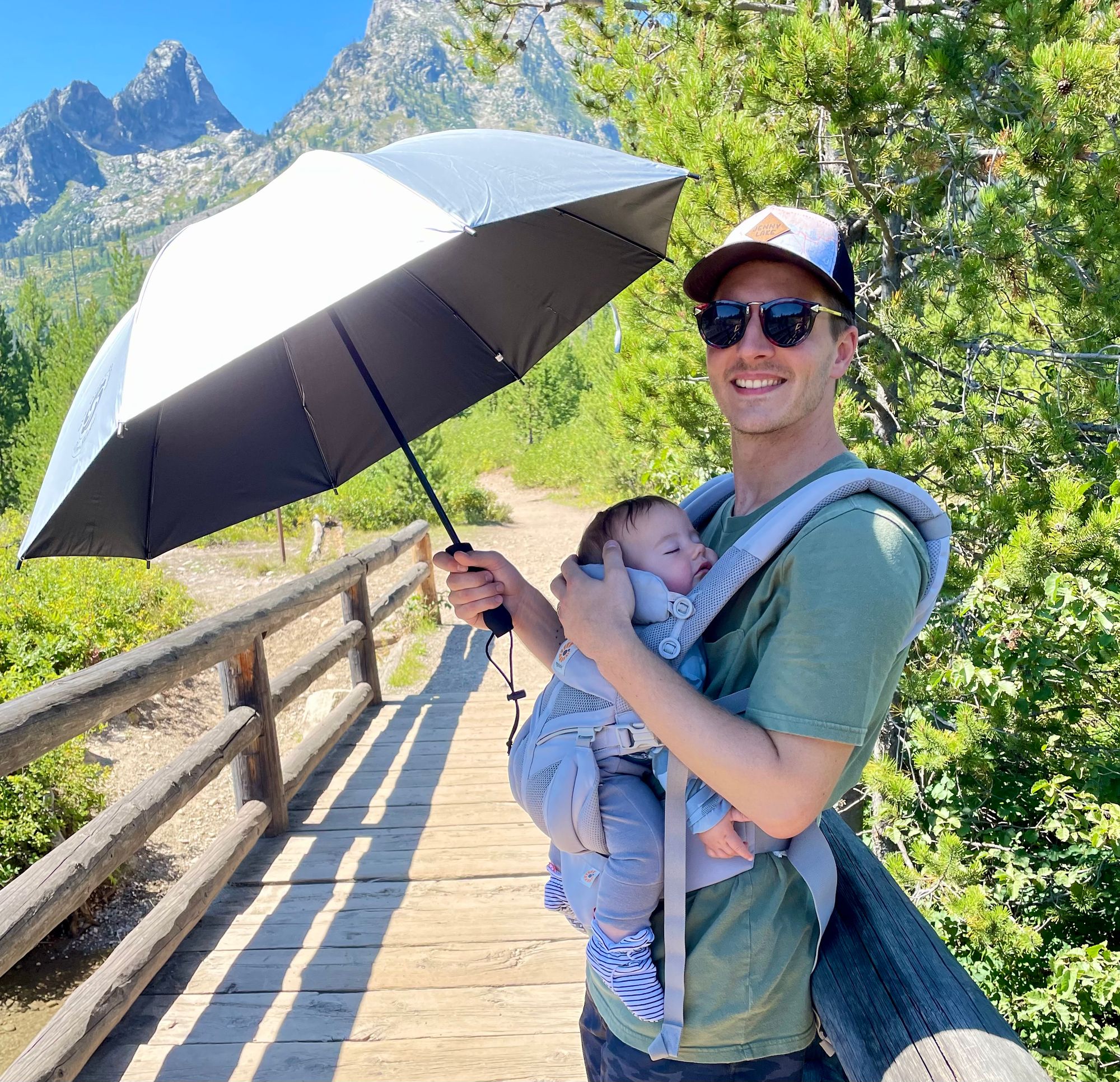 Father baby-wearing and holding sun umbrella while son sleeps in ergobaby carrier